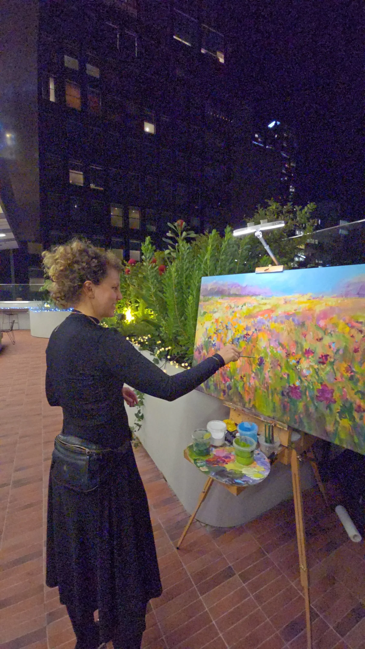 Woman painting an impressionist floral landscape on an easel at night, with plants and a city building in the background.