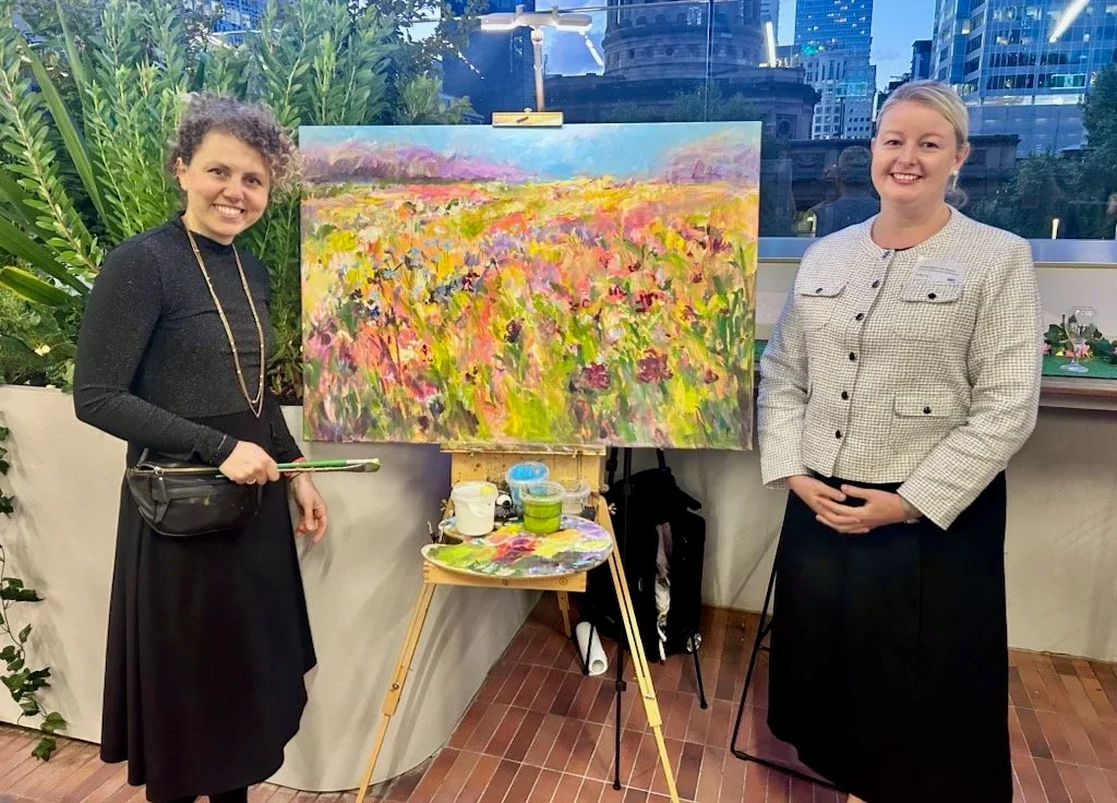 Two women standing next to an easel with a colorful landscape painting inside, surrounded by green plants, in an indoor setting with city buildings visible through the windows.