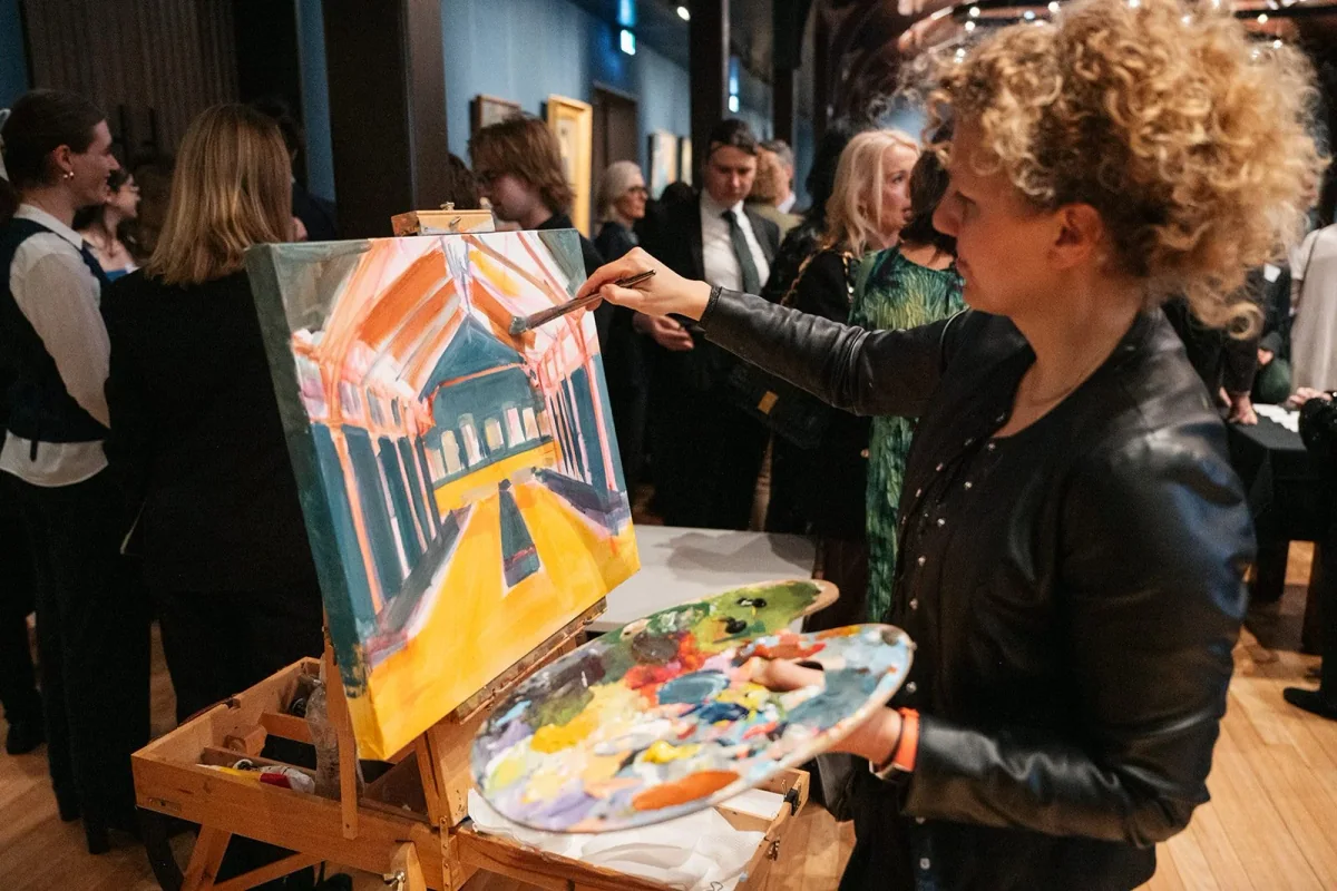 A woman with curly blonde hair wearing a black leather jacket painting a colorful abstract landscape of a building with a large orange roof and yellow ground, surrounded by a crowd of people Trinity College A C C Farran Hall event space.