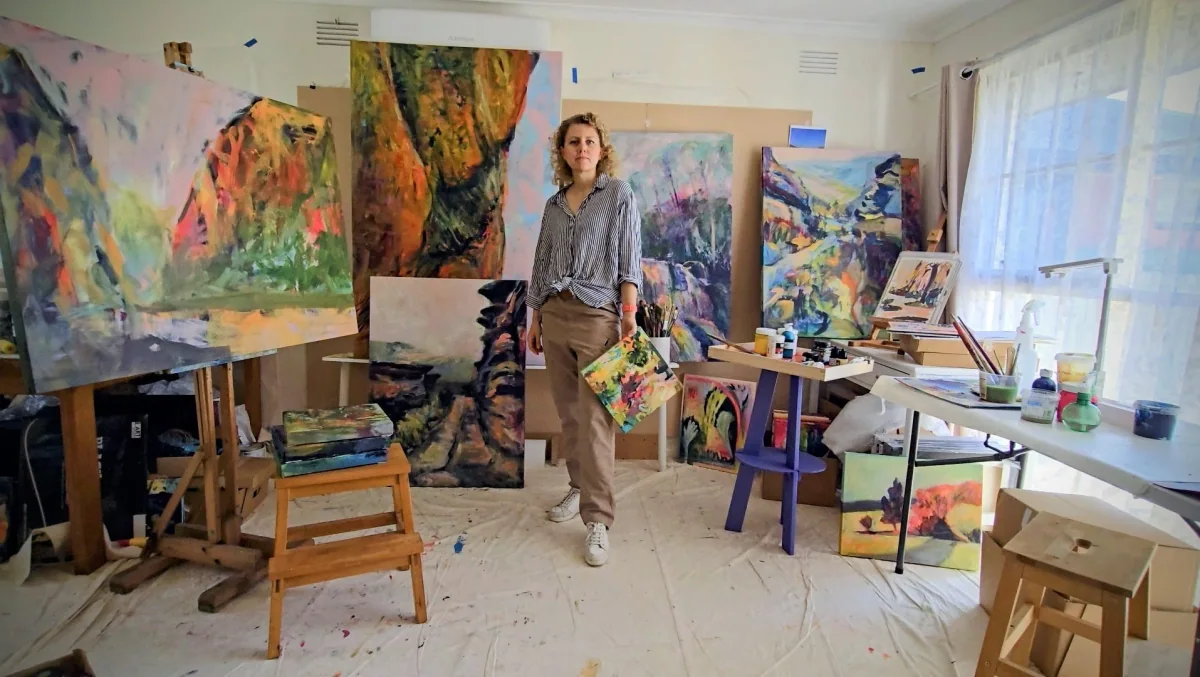 Artist Olga Dziemidowicz standing in a brightly lit studio surrounded by colorful abstract paintings on easels and tables. The studio has large windows with sheer curtains.