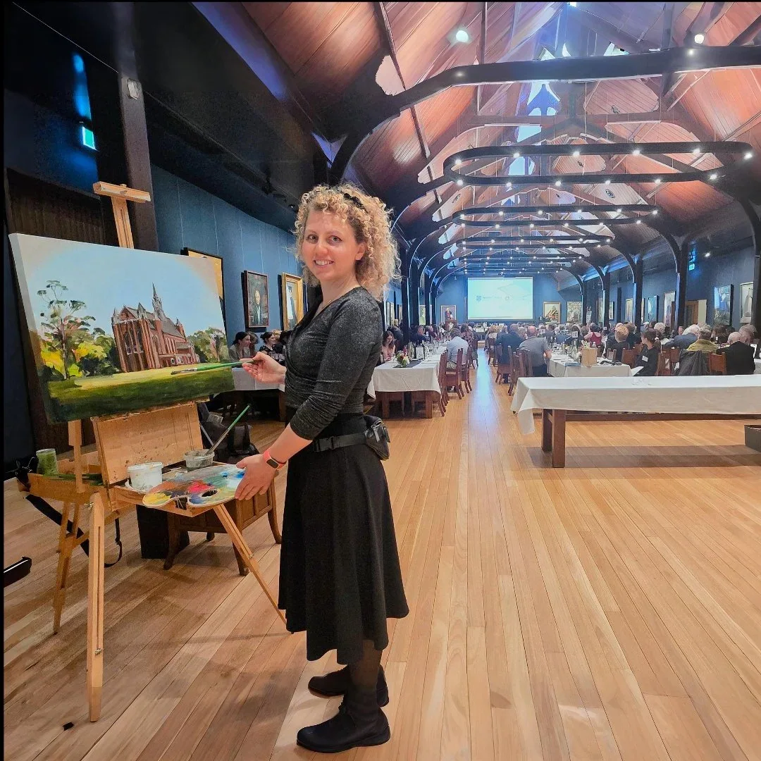 A woman with curly blonde hair standing next to her completed landscape painting on an easel, inside Trinity College with high arched wooden ceilings and a large audience seated at tables in the background.