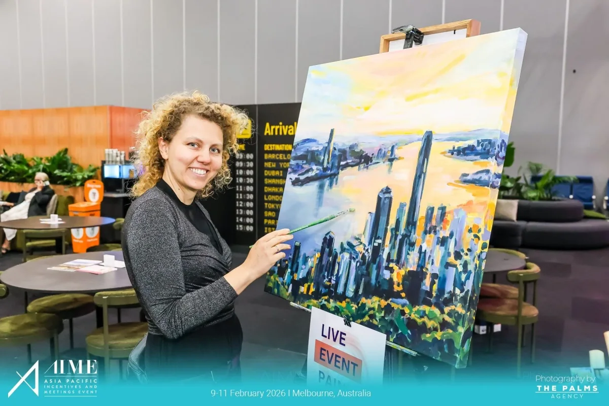 A woman with curly blonde hair smiling while painting a cityscape on a large canvas at an event. The background shows an airport arrival/destination board and lounge area. The image is from the Asia Pacific Incentives and Meetings Event (AIME) held i