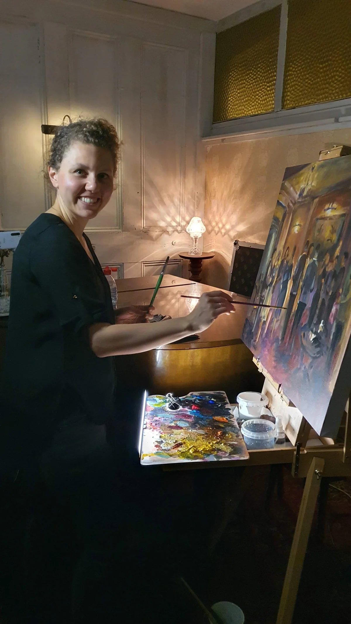 A woman smiling while painting on a canvas in a cozy, warmly lit room with an ornate lamp and art supplies on a table.