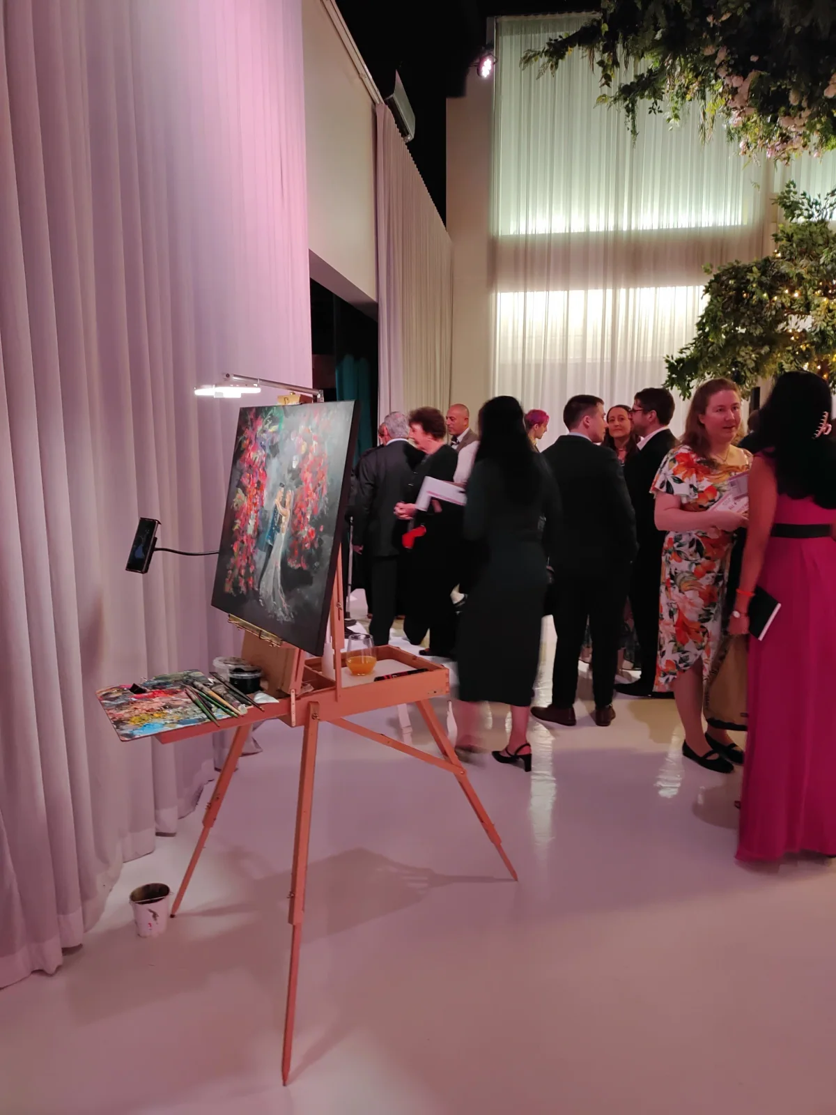 An art gallery event with people socializing, a painting on an easel, and colorful lighting with pink and white curtains in the background.