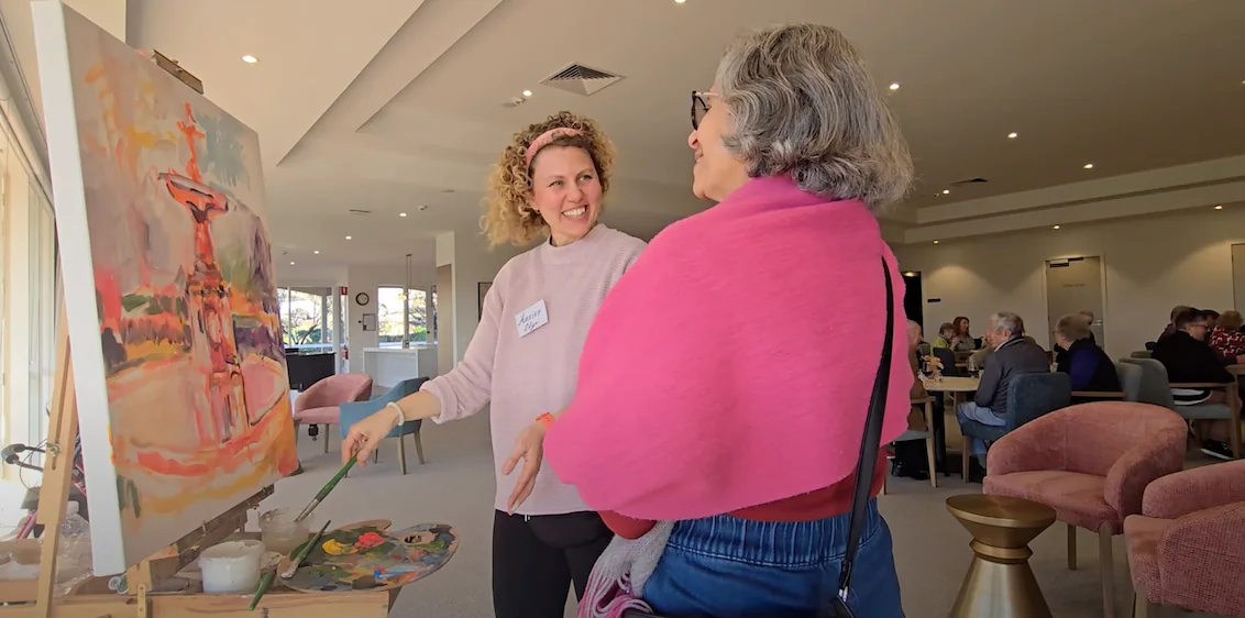 A young woman with curly hair and a pink headband talking with an older woman with gray hair in a pink shawl while live painting
