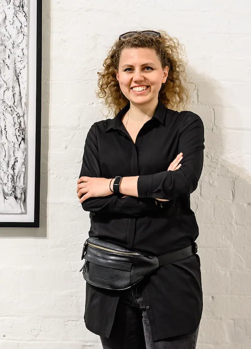 A woman with curly hair smiling and crossing her arms, standing against a white brick wall.