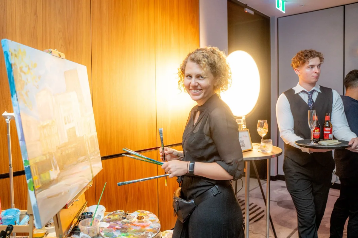 A woman with curly blond hair smiling while holding paintbrushes and standing in front of a canvas on an easel. Two men are in the background, one in a vest and tie holding a tray with drinks. The setting appears to be an indoor social event with woo